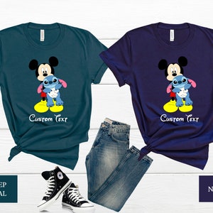 May include: Two t-shirts featuring Mickey Mouse holding Stitch. The shirt on the left is teal and the shirt on the right is navy. Both shirts have the text "Custom Text" printed on them.