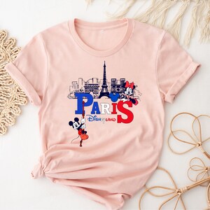 Pu&ograve; includere: Maglietta rosa con una grafica di Topolino e Minnie Mouse di fronte alla Torre Eiffel e il testo "Paris Disneyland".