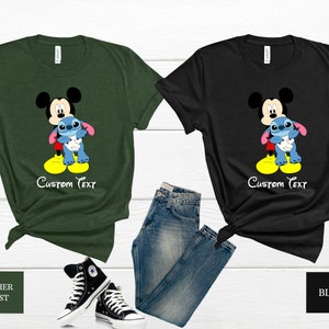 May include: Two t-shirts featuring Mickey Mouse and Stitch. The shirt on the left is a heather forest green colour and the shirt on the right is black. Both shirts have the text "Custom Text" printed on them.