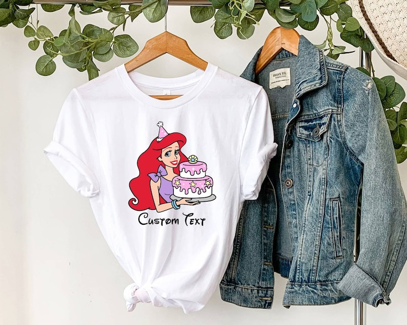 Pode incluir: Camiseta branca com uma imagem de desenho animado de Ariel da Pequena Sereia usando um chap&eacute;u de anivers&aacute;rio e segurando um bolo rosa e branco. O texto "Custom Text" &eacute; impresso abaixo da imagem.