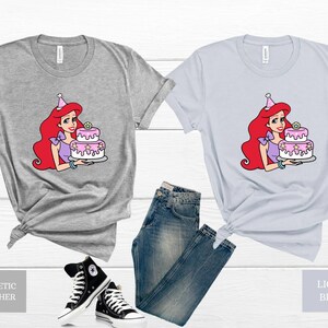 Pode incluir: Duas camisetas com uma imagem de desenho animado de uma mulher de cabelo ruivo usando um chap&eacute;u de festa e segurando um bolo rosa e branco. A camiseta &agrave; esquerda &eacute; cinza heather atl&eacute;tico e a camiseta &agrave; direita &eacute; azul claro.