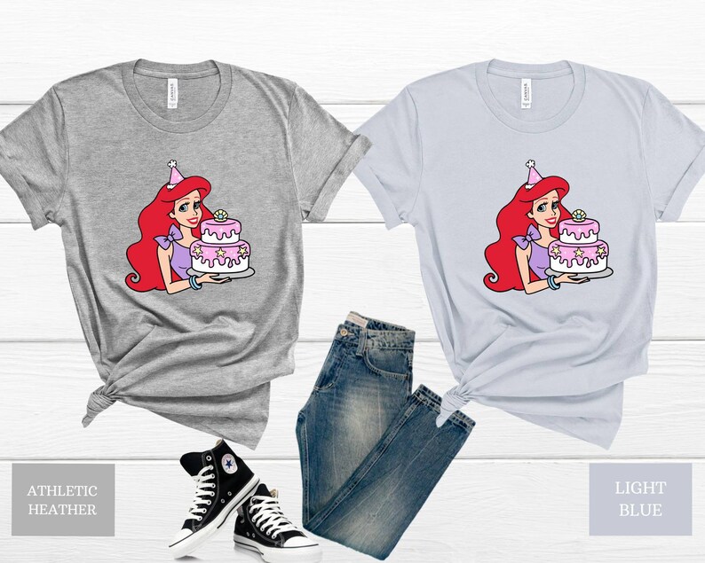 Pode incluir: Duas camisetas com uma imagem de desenho animado de uma mulher de cabelo ruivo usando um chap&eacute;u de festa e segurando um bolo rosa e branco. A camiseta &agrave; esquerda &eacute; cinza heather atl&eacute;tico e a camiseta &agrave; direita &eacute; azul claro.