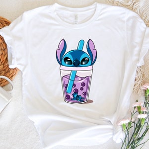 May include: White t-shirt featuring a cartoon illustration of a blue alien character in a purple bubble tea drink. The character has large eyes and large ears. The bubble tea has purple boba pearls and a blue straw.