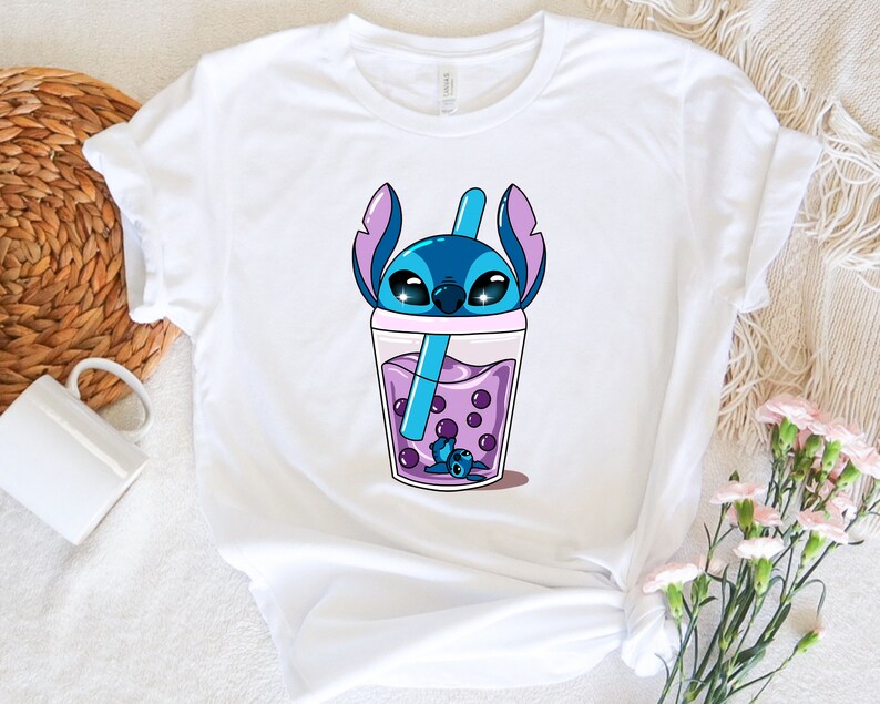 May include: White t-shirt featuring a cartoon illustration of a blue alien character in a purple bubble tea drink. The character has large eyes and large ears. The bubble tea has purple boba pearls and a blue straw.
