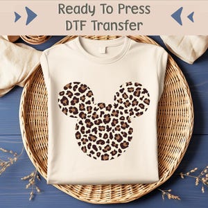 Puede incluir: Una camiseta blanca con un diseño de Mickey Mouse de estampado de leopardo marrón y negro. La camiseta está lista para ser estampada con una transferencia DTF.