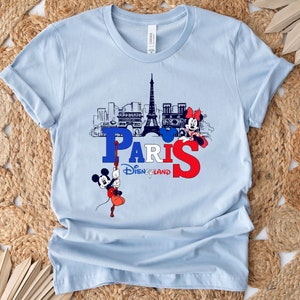 Pu&ograve; includere: Maglietta blu chiaro con una grafica di Topolino e Minnie Mouse di fronte alla Torre Eiffel. La grafica include anche il testo "Paris Disneyland".