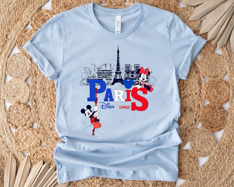 Pu&ograve; includere: Maglietta blu chiaro con una grafica di Topolino e Minnie Mouse di fronte alla Torre Eiffel. La grafica include anche il testo "Paris Disneyland".