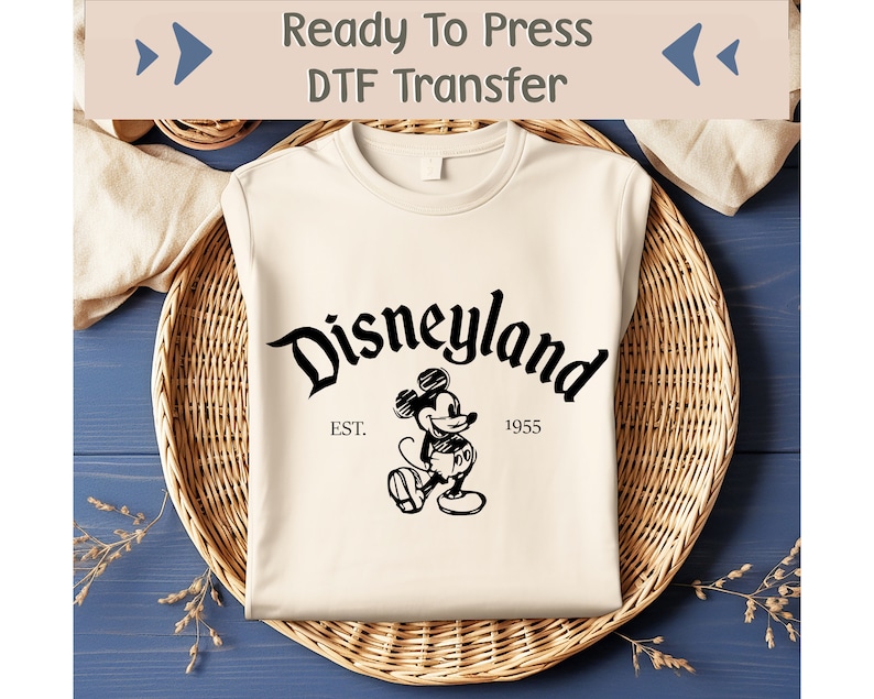 May include: Cream-colored t-shirt with a black Disneyland graphic featuring Mickey Mouse and the text "Disneyland EST. 1955". The shirt is neatly folded in a woven basket.