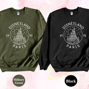 Puede incluir: Dos sudaderas con un dise&ntilde;o gr&aacute;fico blanco del logotipo de Disneyland Par&iacute;s. El logotipo representa un dibujo estilizado del castillo de la Bella Durmiente con el texto "Disneyland Par&iacute;s" y "1992" alrededor del castillo. La sudadera de la izquierda es verde militar y la sudadera de la derecha es negra.
