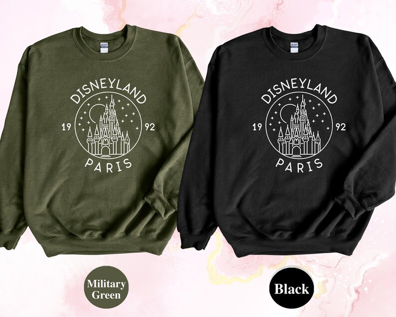 Puede incluir: Dos sudaderas con un dise&ntilde;o gr&aacute;fico blanco del logotipo de Disneyland Par&iacute;s. El logotipo representa un dibujo estilizado del castillo de la Bella Durmiente con el texto "Disneyland Par&iacute;s" y "1992" alrededor del castillo. La sudadera de la izquierda es verde militar y la sudadera de la derecha es negra.