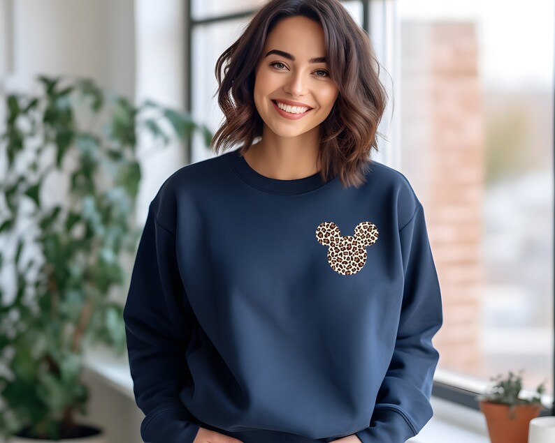 May include: Navy blue sweatshirt featuring a leopard print Mickey Mouse design on the chest. The sweatshirt has a crew neck and long sleeves. The Mickey Mouse design is made up of brown and tan spots. The background is a solid navy blue.