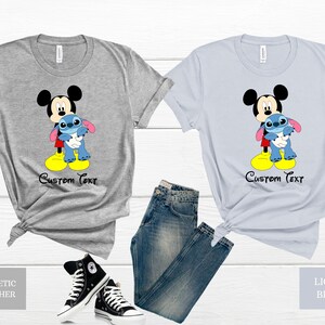 May include: Two t-shirts featuring Mickey Mouse holding Stitch. The shirt on the left is athletic heather grey and the shirt on the right is light blue. Both shirts have the text "Custom Text" printed below the image.