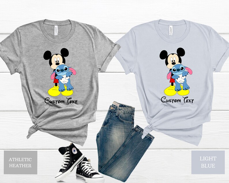 May include: Two t-shirts featuring Mickey Mouse holding Stitch. The shirt on the left is athletic heather grey and the shirt on the right is light blue. Both shirts have the text "Custom Text" printed below the image.