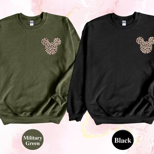 May include: Two crewneck sweatshirts, one military green and one black. Each features a leopard print Mickey Mouse head design on the upper left chest. The military green sweatshirt has the words "Military Green" in a circle below it, and the black sweatshirt has the word "Black" in a circle below it.