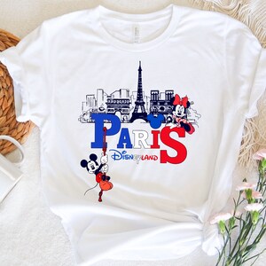Pu&ograve; includere: Maglietta bianca con una grafica di Topolino e Minnie Mouse di fronte alla Torre Eiffel e la parola "Parigi" con il testo "Disneyland" sotto.