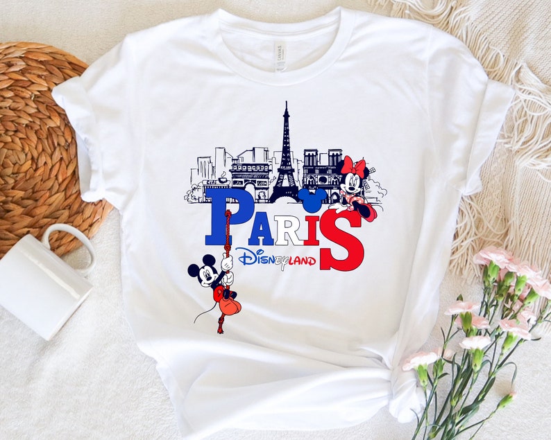 Pu&ograve; includere: Maglietta bianca con una grafica di Topolino e Minnie Mouse di fronte alla Torre Eiffel e la parola "Parigi" con il testo "Disneyland" sotto.