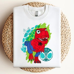 May include: White t-shirt with a red and blue dinosaur wearing a Spiderman costume. The dinosaur is standing on a green and blue background with a white spider web.