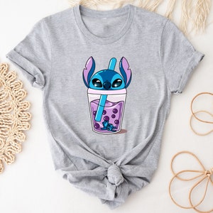 May include: A light gray t-shirt featuring a cartoon character in a boba drink. The character is blue with large eyes and is inside a clear cup with purple boba pearls and a blue straw. The shirt has short sleeves and a crew neck.