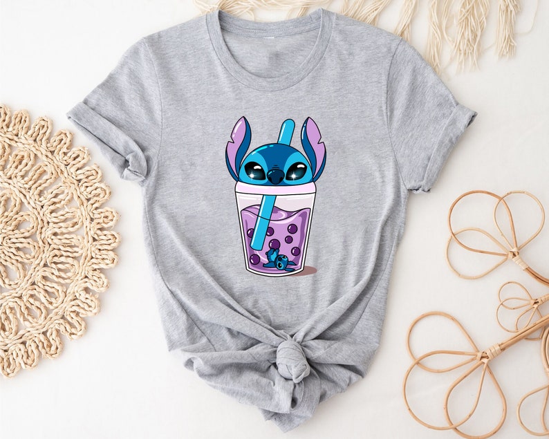 May include: A light gray t-shirt featuring a cartoon character in a boba drink. The character is blue with large eyes and is inside a clear cup with purple boba pearls and a blue straw. The shirt has short sleeves and a crew neck.
