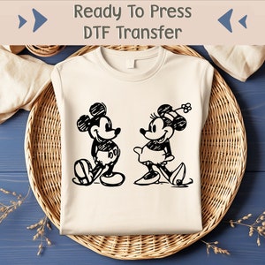 May include: A white t-shirt featuring a black and white image of Mickey Mouse and Minnie Mouse. The text "Ready To Press DTF Transfer" is at the top of the image.