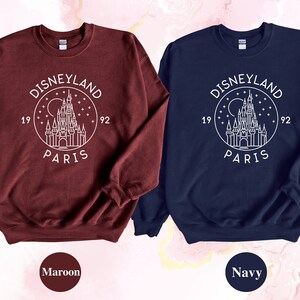 Puede incluir: Dos sudaderas con un contorno blanco del castillo de Disneyland Par&iacute;s y el texto "DISNEYLAND PARIS 1992" en un c&iacute;rculo. La sudadera de la izquierda es granate y la sudadera de la derecha es azul marino.