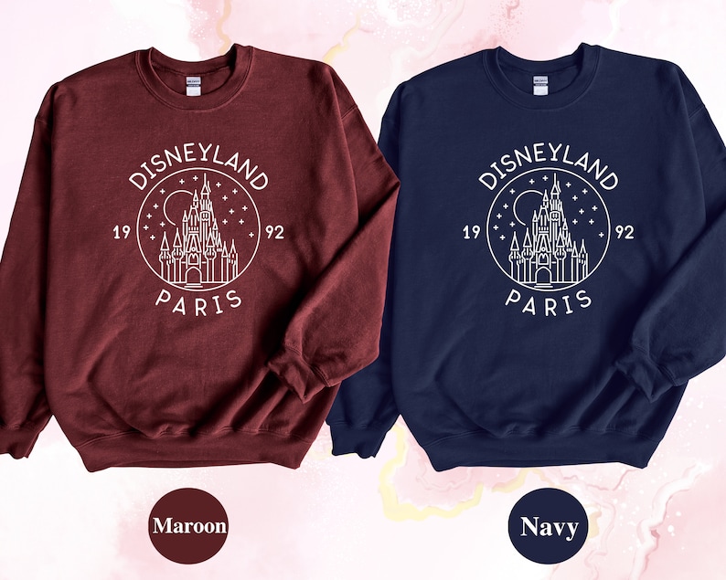 Puede incluir: Dos sudaderas con un contorno blanco del castillo de Disneyland Par&iacute;s y el texto "DISNEYLAND PARIS 1992" en un c&iacute;rculo. La sudadera de la izquierda es granate y la sudadera de la derecha es azul marino.