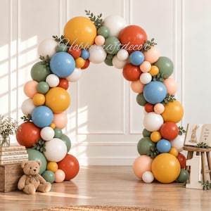 May include: A large balloon arch featuring a mix of matte balloons in mustard yellow, terracotta orange, sage green, dusty blue, cream, and soft peach, accented with small green foliage sprigs throughout the structure for a decorative display.