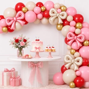 May include: A balloon arch featuring red, pink, beige, and gold balloons with pink and beige foil bow balloons. A cake with cherries, cupcakes, a flower bouquet, macarons, and two gift boxes with pink ribbons are arranged on a white table.