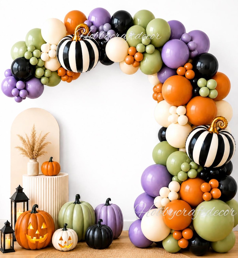 May include: A balloon arch featuring orange, purple, black, olive green, and cream balloons with two black and white striped pumpkin balloons. Below are several decorative pumpkins in orange, green, purple, and black, including two lit jack-o'-lanterns.