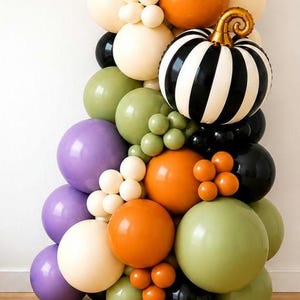 May include: A balloon garland featuring a mix of round balloons in orange, cream, sage green, purple, and black. A large, round, black and white striped foil balloon shaped like a pumpkin with a metallic gold stem is integrated into the arrangement.