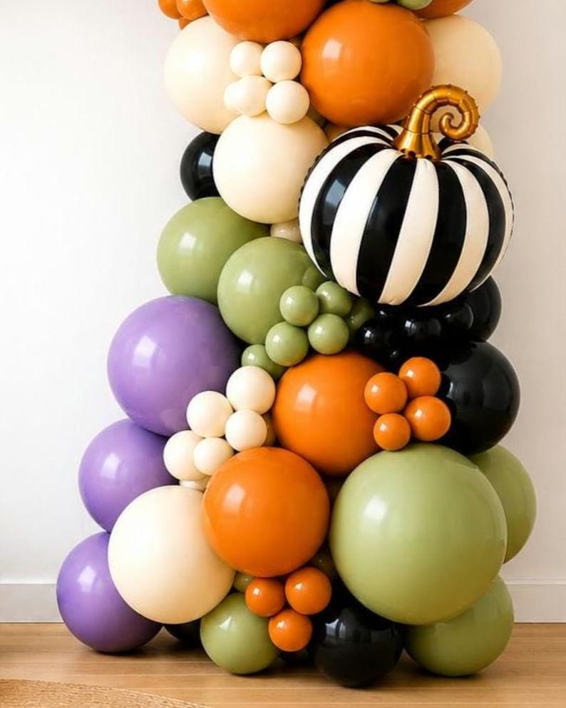 May include: A balloon garland featuring a mix of round balloons in orange, cream, sage green, purple, and black. A large, round, black and white striped foil balloon shaped like a pumpkin with a metallic gold stem is integrated into the arrangement.