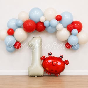 May include: A balloon arch featuring a mix of light blue, white, and red balloons in various sizes. Below the arch stands a large, cream-colored number one balloon and a red crab-shaped balloon with a smiling face and pink cheeks.