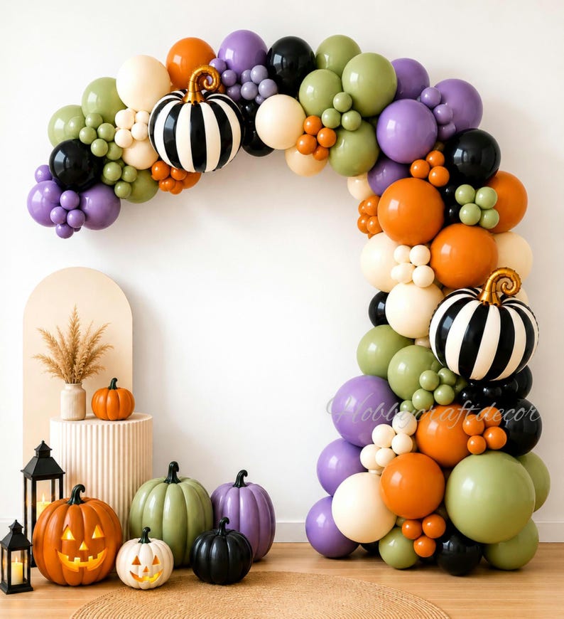 May include: A balloon arch featuring orange, purple, olive green, black, and cream balloons with two black and white striped pumpkin balloons. Below, several decorative pumpkins in orange, green, purple, and black sit near two black lanterns.