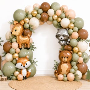 May include: A balloon arch featuring sage green, brown, cream, and gold balloons with foil animal balloons shaped like a deer, raccoon, fox, and bear. A wooden signpost reads BE BRAVE, BE KIND, EXPLORE, and a small sign reads Let the adventure BEGIN.