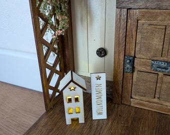 May include: A miniature wooden house with a glowing yellow interior and a star cutout on the roof, positioned next to a rectangular wooden sign with the word WILLKOMMEN and a star symbol, set against a dark wood structure with a trellis.
