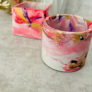 Puede incluir: Dos pequeñas macetas de hormigón pintadas a mano. Una es cuadrada, la otra cilíndrica. Ambas presentan un diseño veteado con tonos rosas, blancos, amarillos y morados. Las macetas son adecuadas para plantas pequeñas o suculentas.