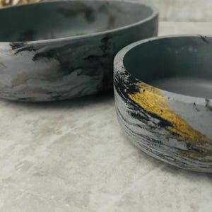 May include: Two gray and gold decorative bowls. The bowls have a marbled effect with black and gold accents. The bowls are different sizes and have a smooth, rounded shape.