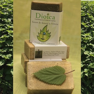 May include: A stack of three green and brown bars of soap with a white label that reads "Dioica Cosmétiques à l'ortie de Sarthe Savon de beauté à l'Ortie VISAGE-CORPS-CHEVEUX Ingrédients: Sodium Olivate, Aqua, Glycerin, Olea Europaea Fruit Oil, Urtica Dioica Leaf Powder". A green leaf is on top of the soap.