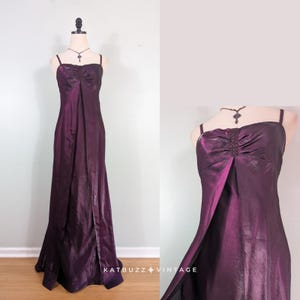 May include: A floor-length, deep purple iridescent gown featuring thin spaghetti straps, a ruched bodice with a central beaded vertical accent, and a straight skirt with a front slit. The dress is displayed on a mannequin with a necklace.