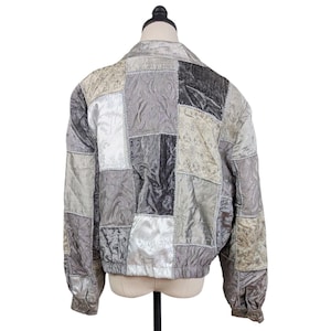 May include: A long-sleeve jacket featuring a patchwork design composed of various shades of gray, silver, and white fabric panels. The garment displays diverse textures and subtle floral patterns, with visible stitching connecting each square section.