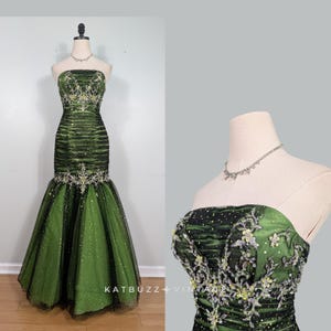 Peut inclure: Une robe longue sans bretelles dans des tons vert foncé et noir. Le corsage présente des fronces horizontales, des broderies florales argentées et jaunes complexes, ainsi que des sequins. La jupe s'évase en une silhouette sirène en tulle pailleté.