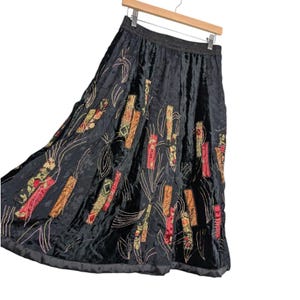 May include: Black velvet skirt with colorful embroidered accents. The skirt features a flowing A-line silhouette and is adorned with vertical rectangular and leaf-shaped embroidered patches in shades of red, orange, and green. The skirt hangs from a wooden hanger.