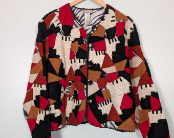 May include: A long-sleeve jacket featuring a geometric patchwork design in red, black, tan, and cream. The garment has a round neckline, a front button closure with five black buttons, and a textured fabric construction with varied shapes.