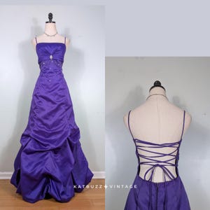 Puede incluir: Un vestido largo de color morado con escote recto, tirantes finos y un corpiño fruncido con un adorno decorativo central. La falda tiene un diseño drapeado y la parte posterior presenta un cierre de corsé con cordones finos.