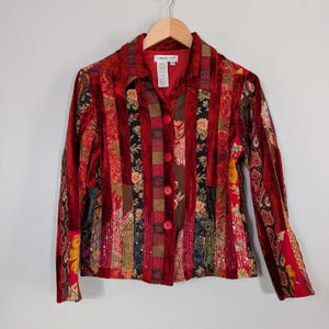 May include: A red velvet jacket with a patchwork design. The jacket features vertical stripes of floral, paisley, and sequined fabrics. It has a collared neckline, long sleeves, and red buttons down the front. The label reads "Coldwater Creek".