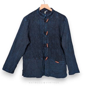 May include: A dark blue jacket with a mandarin collar and wooden toggle closures. The jacket has long sleeves and two front pockets. The fabric has a textured appearance. The jacket is hanging on a wooden hanger. The text "KATBUZZ VINTAGE" is at the bottom.