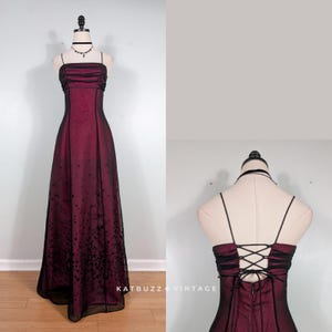 Könnte beinhalten: Ein bodenlanges Kleid mit einer tief magentafarbenen Basisschicht und einem schwarzen, transparenten Überzug. Das Oberteil hat horizontale Raffungen und dünne Spaghettiträger. Der Rock zeigt ein schwarzes Sternenmuster, das zum Saum hin verblasst. Der Rücken verfügt über eine Korsettschnürung mit dünnen schwarzen Bändern.