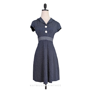 Puede incluir: Un vestido azul marino con estampado de lunares blancos, mangas cortas, cuello y dos botones blancos grandes en el corpiño. Una banda horizontal texturizada se sitúa en la cintura y la falda se ensancha ligeramente hacia el dobladillo.