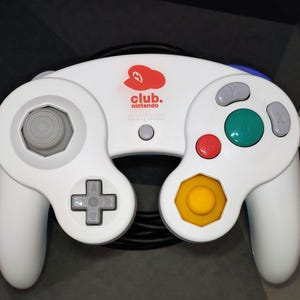 May include: White Nintendo GameCube controller with a Club Nintendo logo. Features a gray joystick, directional pad, and buttons in red, green, and yellow. Includes a black cable and a plug.