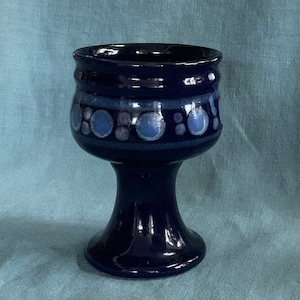 Puede incluir: Un cáliz de cerámica azul oscuro con un borde ancho y una base de pedestal. El cáliz presenta una banda de círculos azules claros y blancos alrededor del centro. El interior del cáliz es de un tono azul más oscuro.